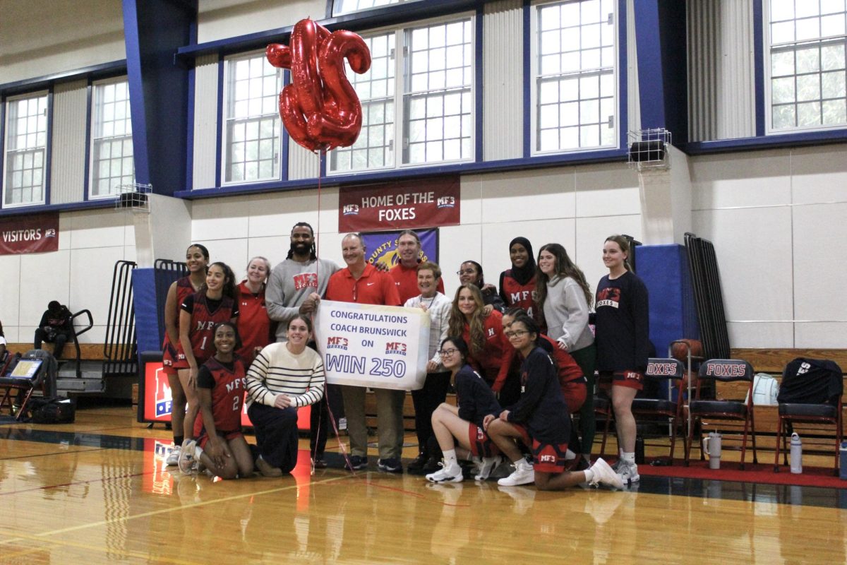 Michael Brunswick, Isaiah Coleman, Debra Williams, and the Varsity Girls’ Basketball Team pose for a photo with a sign and balloons. The sign reads, “Congratulations Coach Brunswick on Win 250,” while the balloons spell out “250.” Photo by Charlotte Nesevich ’25.